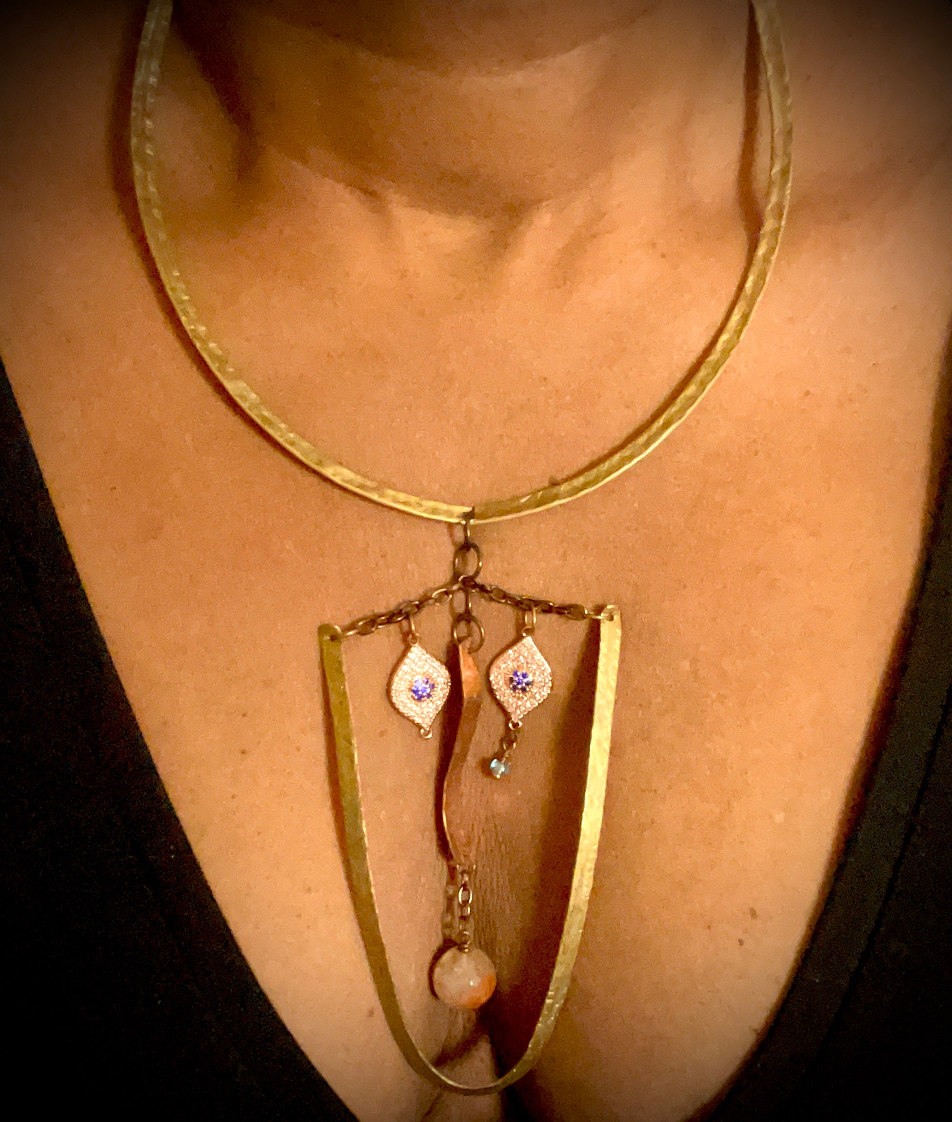 Close up of a woman’s neck and cleavage area shown wearing a brass collar necklace with elongated face made of hammered brass, eyes made of evil eye charms, nose made of hammered, curved copper suspended in middle from a length of chain. Copper has a round agate bead hanging to make the mouth.