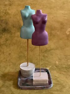 Photo in front of moss green backdrop of two miniature mannequin torsos on brass wire with square or concrete base support. One mannequin is green and one is purple and they are small enough to fit atop a drink coaster, which is also illustrated.