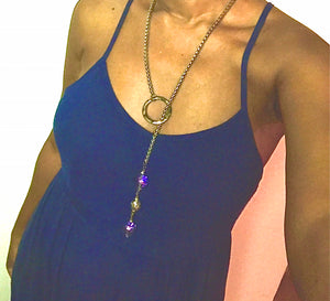 Photo of woman's torso in blue spaghetti strap dress showing off lariat style necklace made of  thick stainless steel chain and a distressed metal circle through which two chunky, iridescent crystal beads and a metal bead dangle.