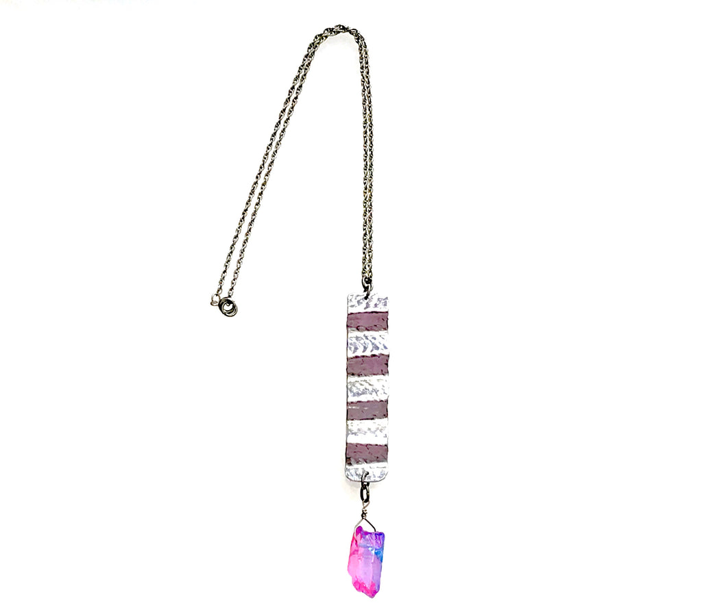 Shows a necklace on a chain with a pendant made from hammered and fold-formed aluminum. A pink and bluish colored crystal hangs from the elongated piece of hammered, fold-formed aluminum.