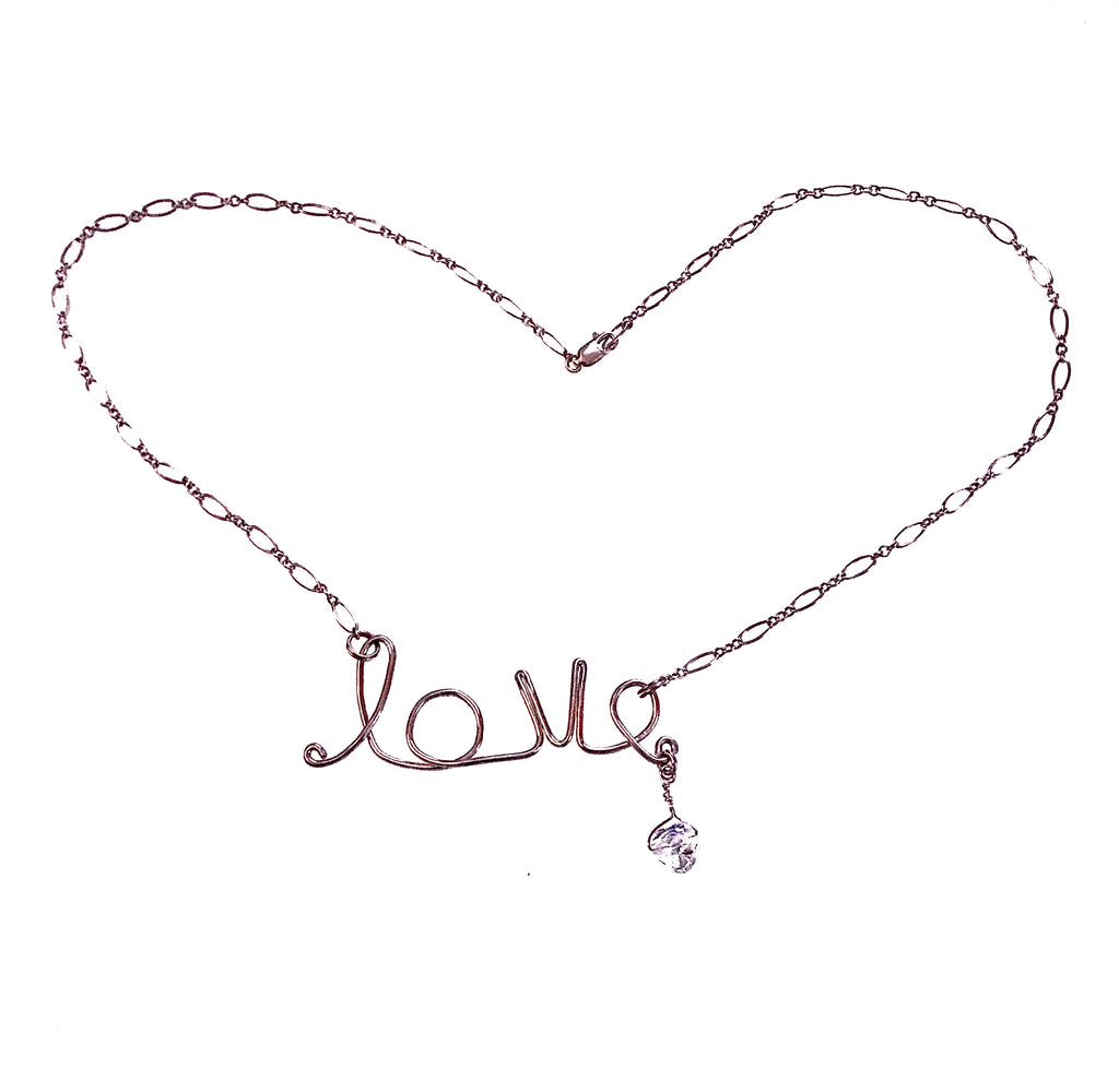 Photo of a sterling silver chain necklace with the word love in the middle (horizontally) as a pendant. The love is hand forged from silver wire. A heart-shaped Swarovski crystal dangles from the e in love. 